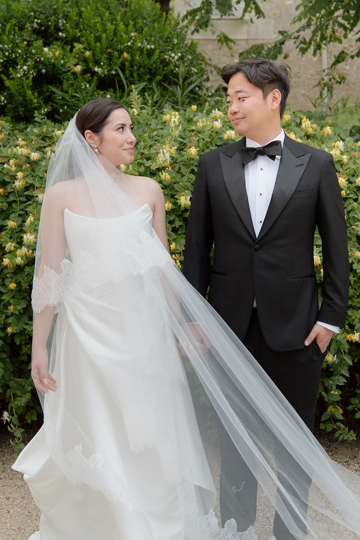 Carina and Stefan standing in front of a flowering hedge, her lace-trimmed veil trailing