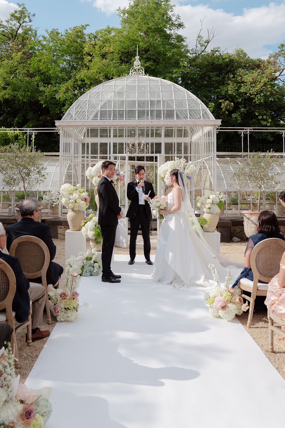 Carina and Stefan with the officiant at the head of the aisle in front of the glass greenhouse