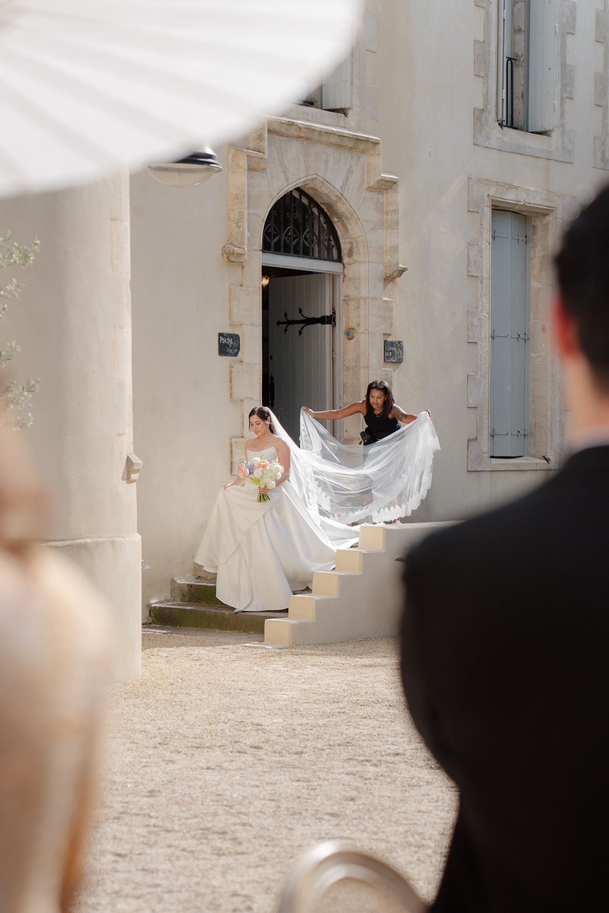 Carina stepping down the château steps as a woman spreads out her long veil
