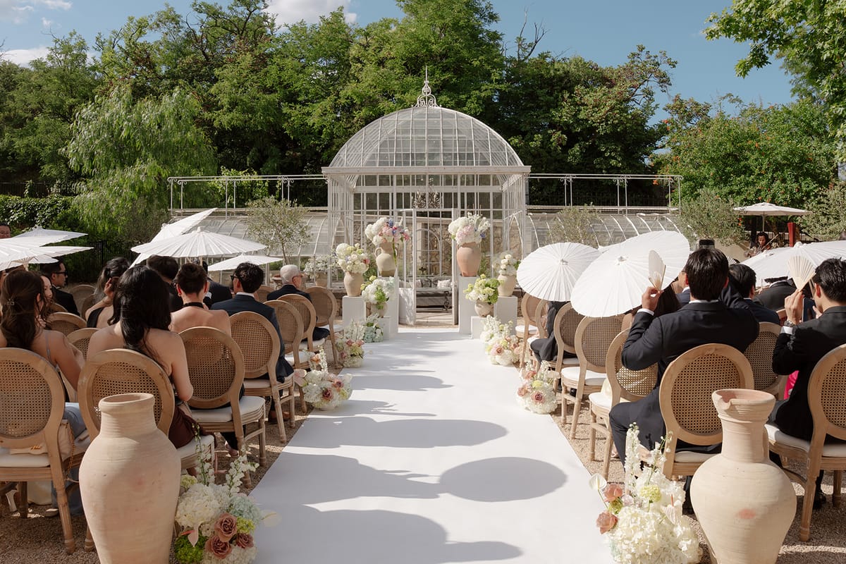 View along the aisle to the glass greenhouse, guests holding white paper parasols