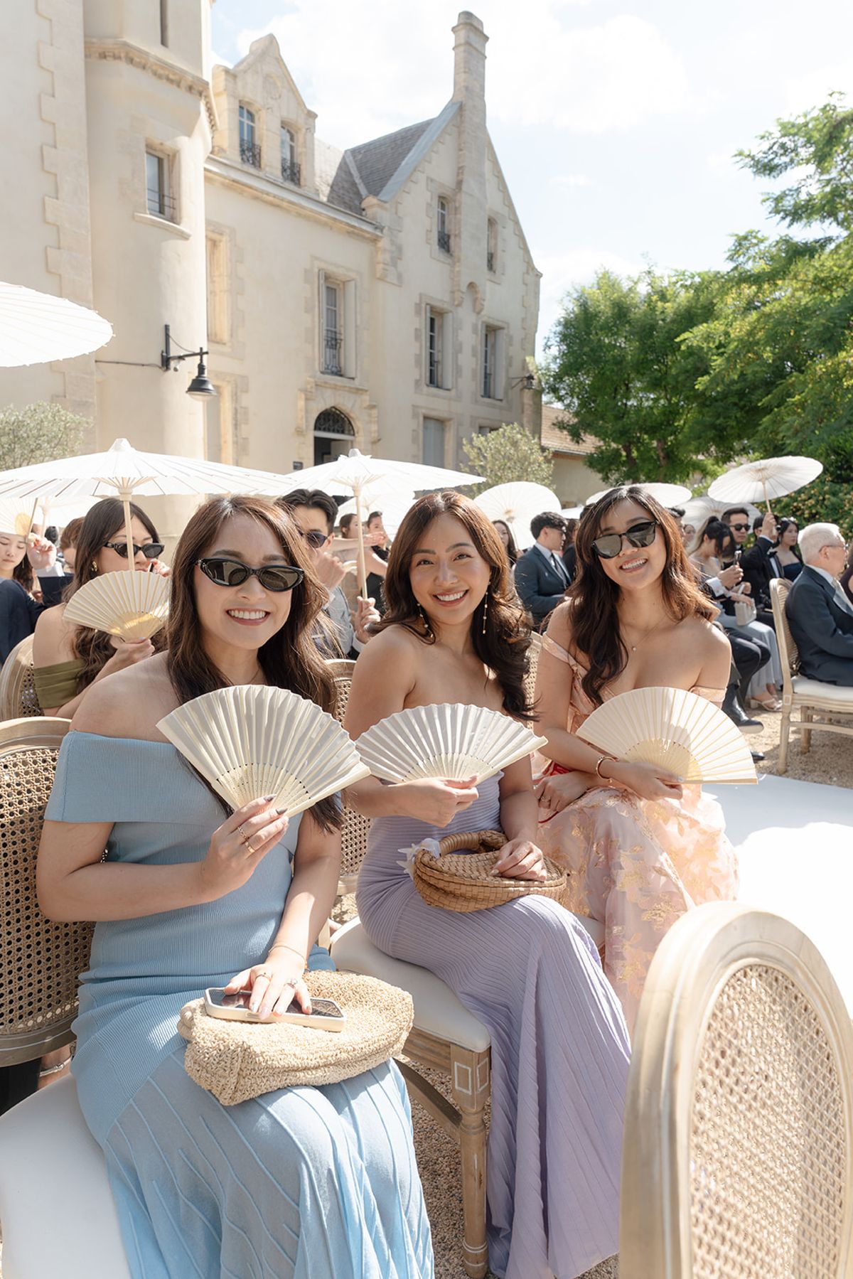 Three guests in pastel dresses and sunglasses seated with paper fans before the ceremony