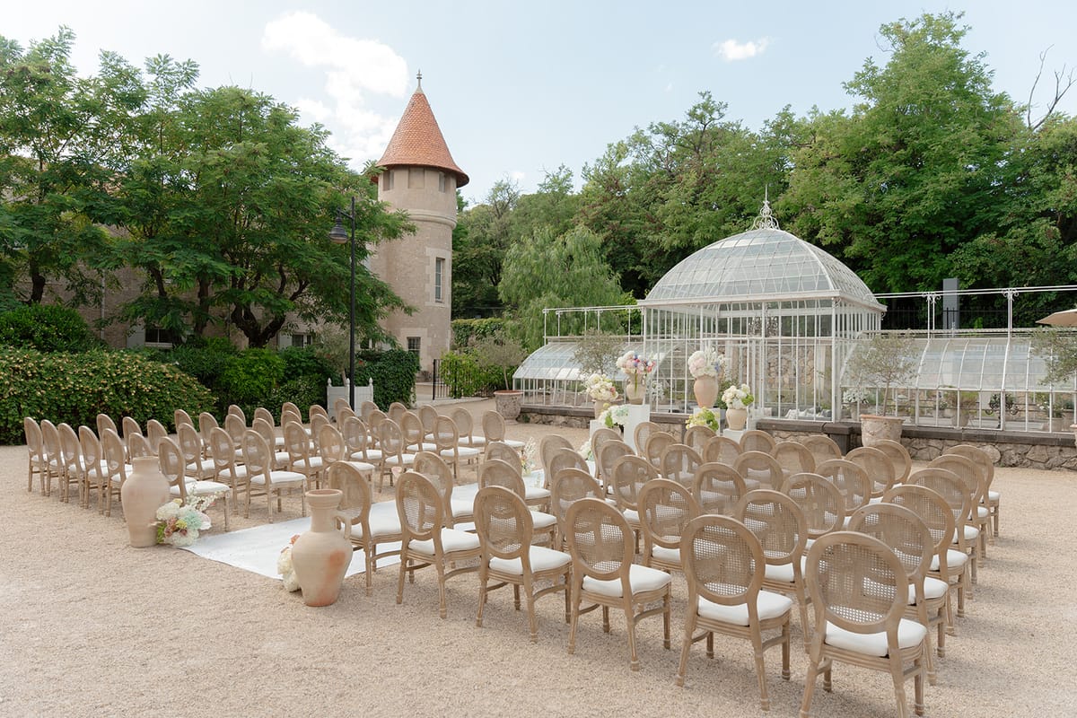 Wide view of the ceremony seating before the glass greenhouse, a turret among the trees