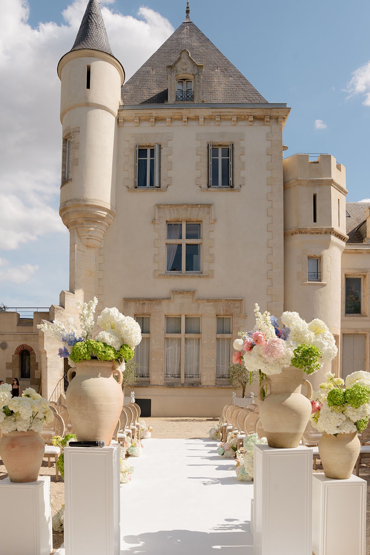 Urns of pastel flowers on white pedestals lining an aisle towards the façade of Château les Carrasses