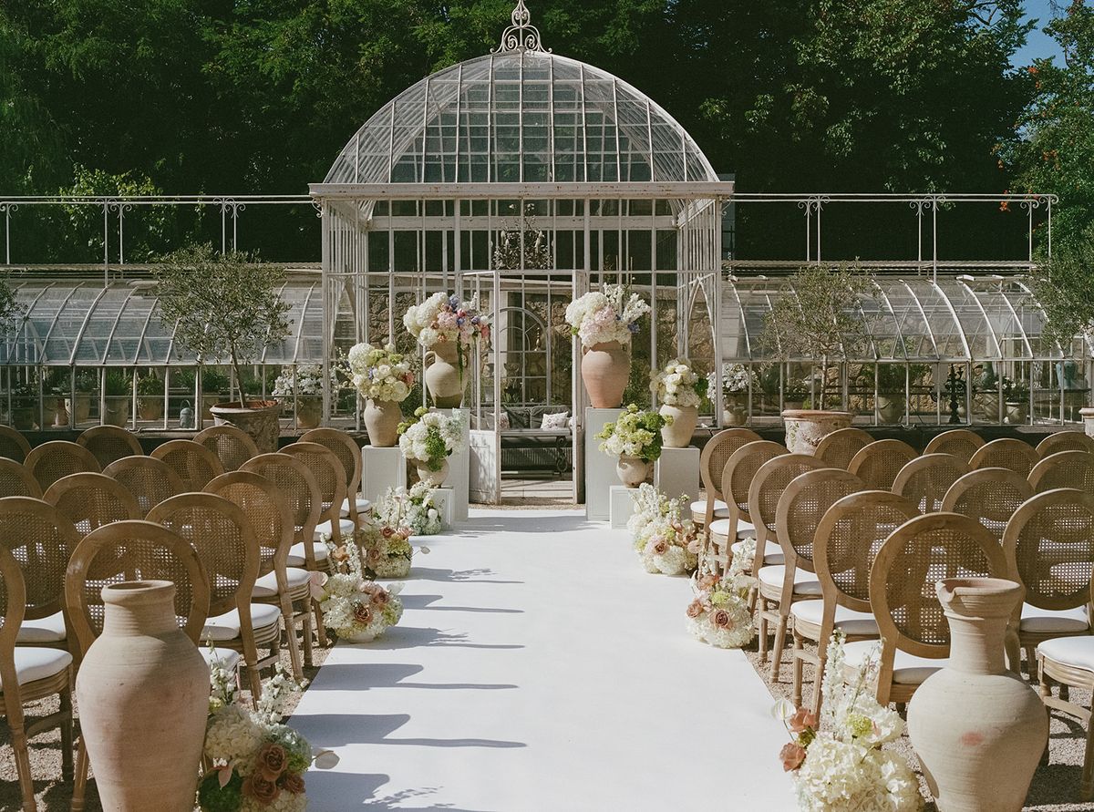The ceremony aisle with terracotta urns and pastel florals leading to the glass greenhouse