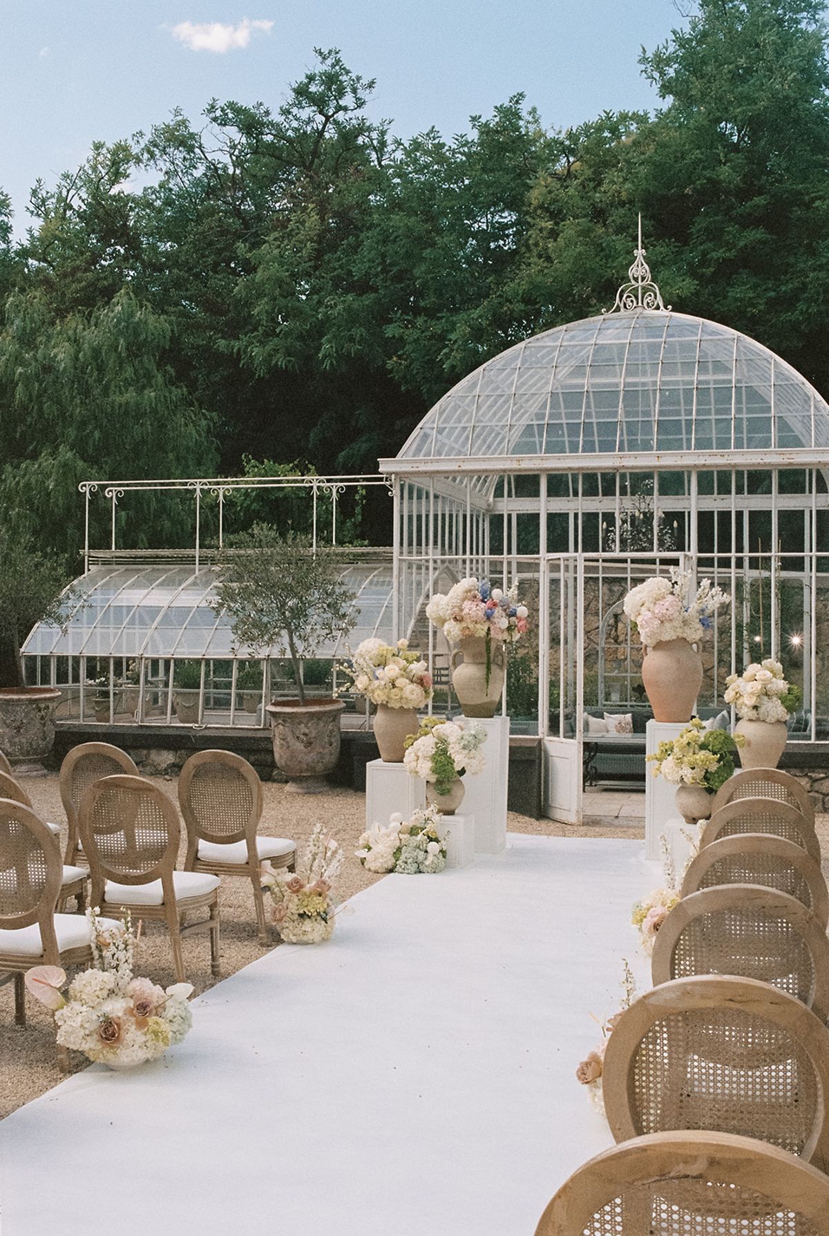 Cane-back chairs and pastel floral arrangements along the aisle in front of the glass greenhouse