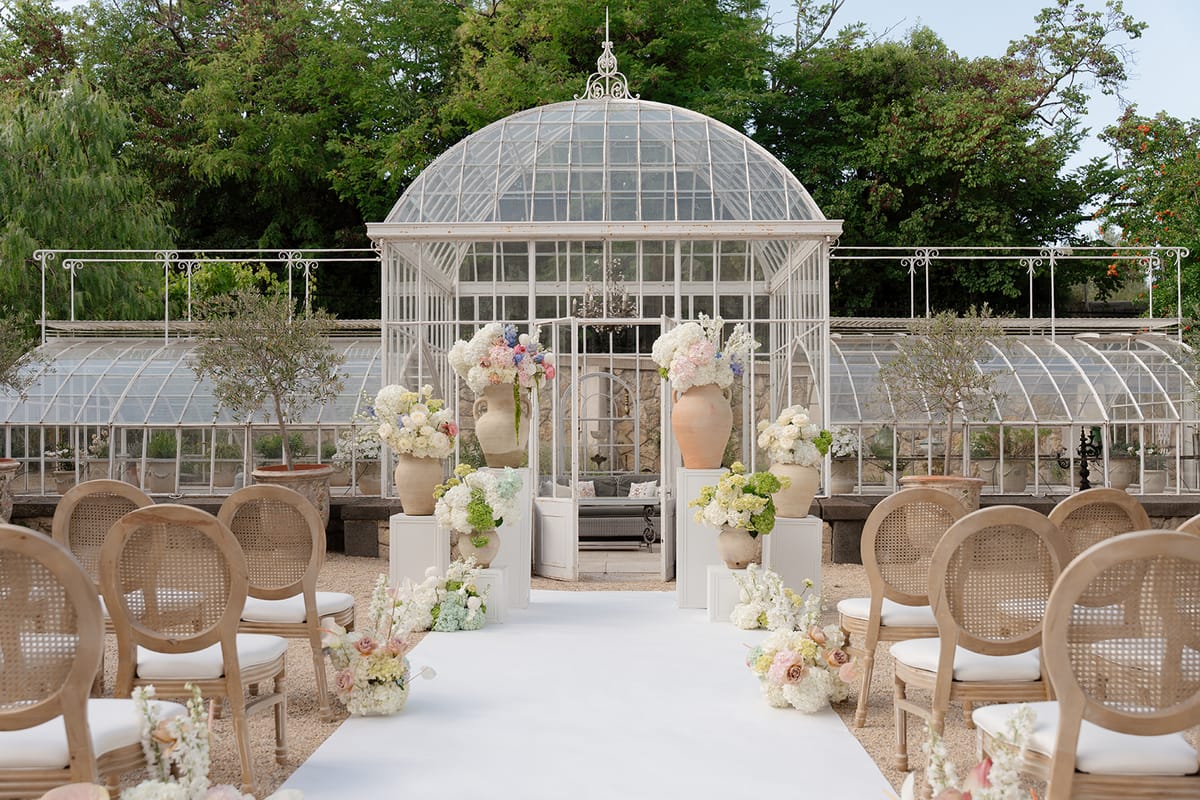 Ceremony aisle in white leading to the glass greenhouse, lined with cane-back chairs and urns of flowers