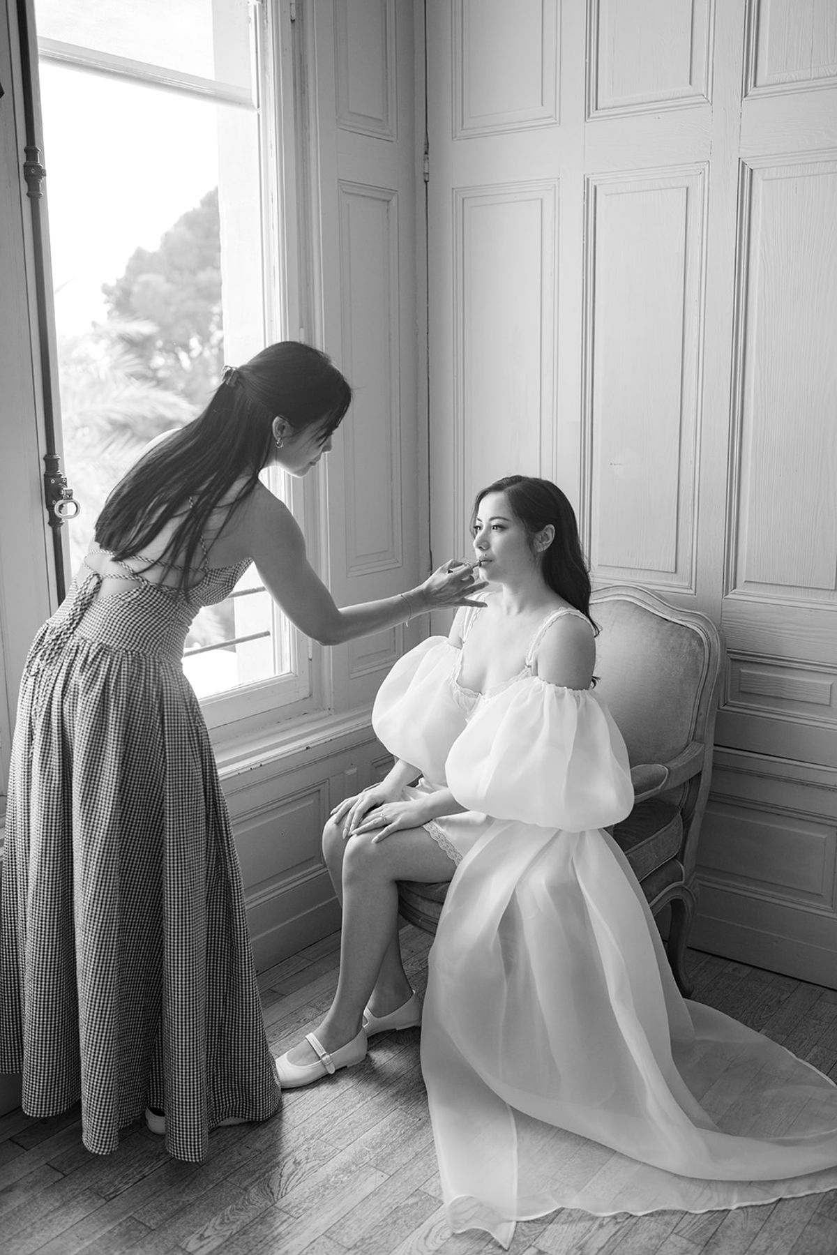 Black and white view of a makeup artist working on Carina, seated by a tall window