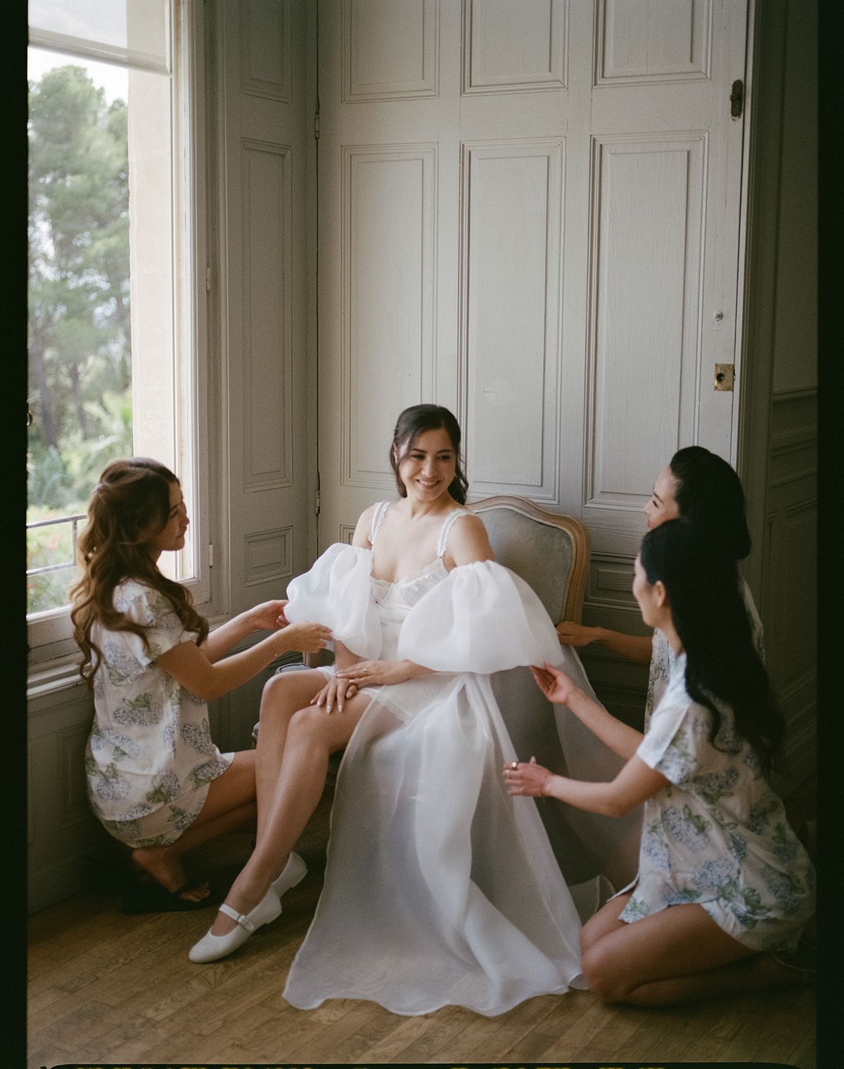 Carina seated in a white robe cape while three bridesmaids in matching floral pyjamas adjust it