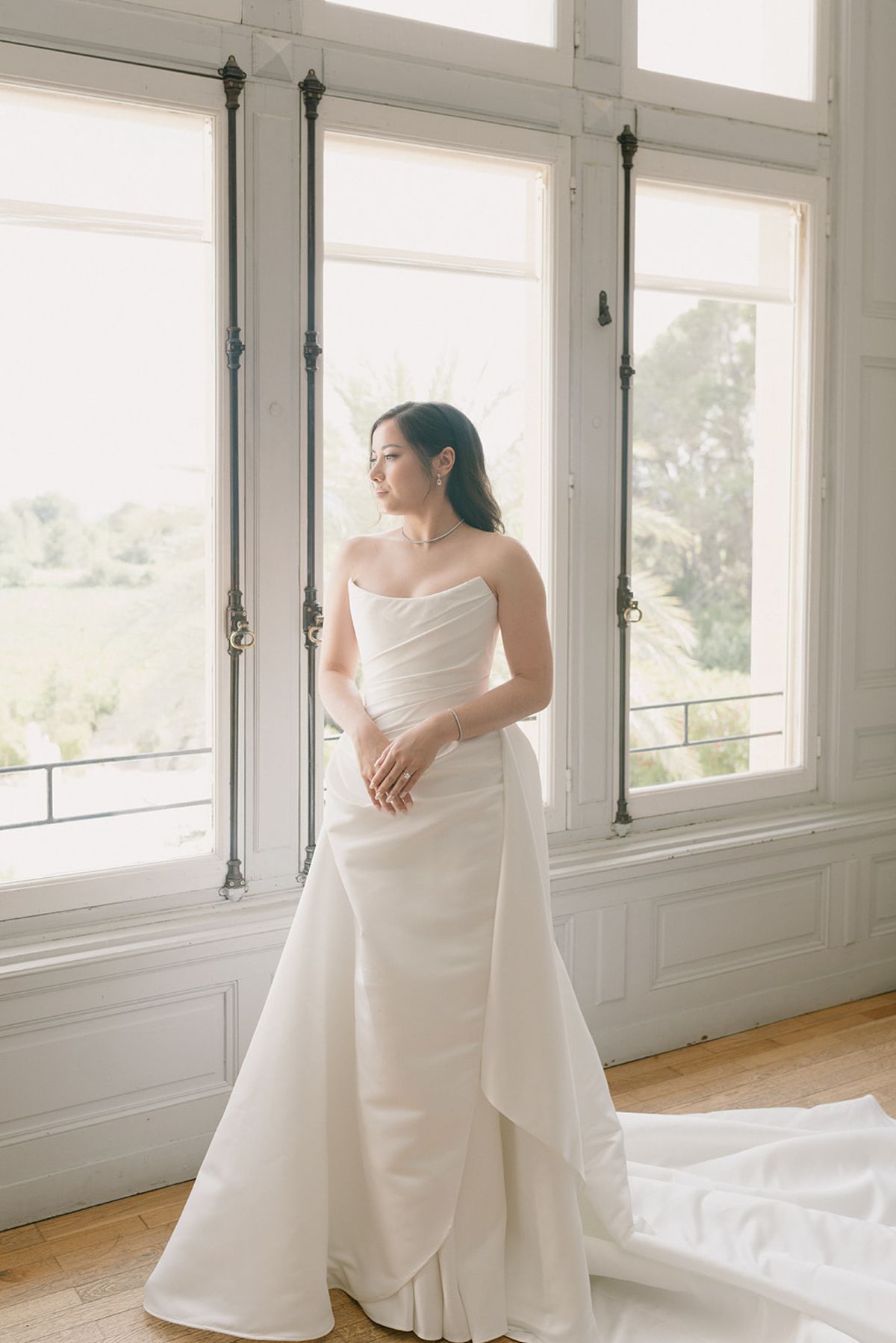 Carina in her strapless ivory gown standing by tall windows in the château