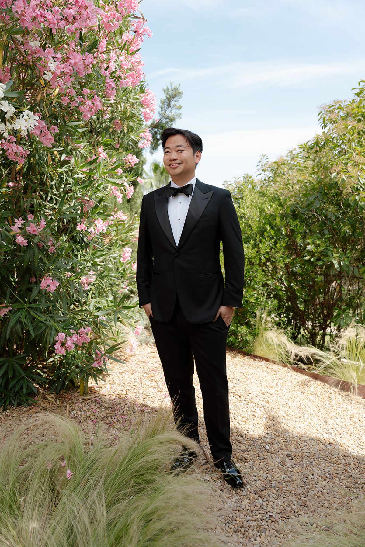 Stefan in a black tuxedo standing on a gravel path beside pink oleander