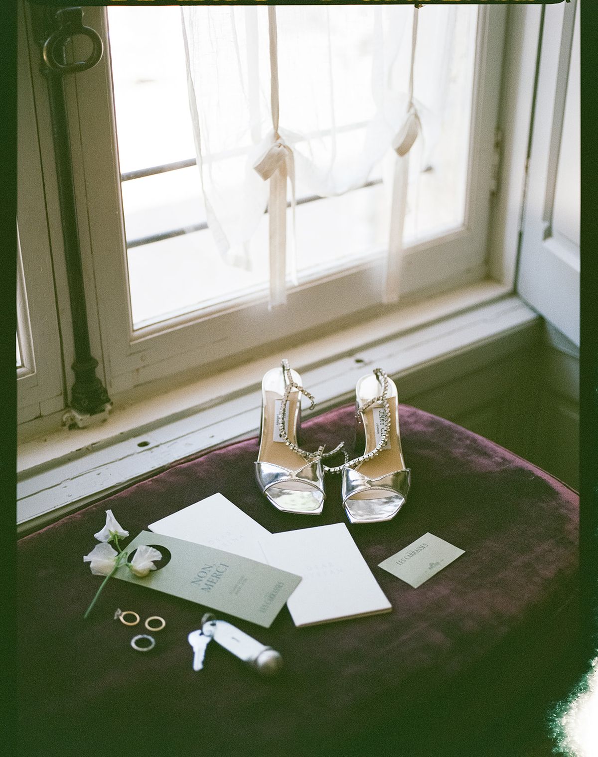Silver heels, wedding rings and stationery arranged on a velvet chair by a window