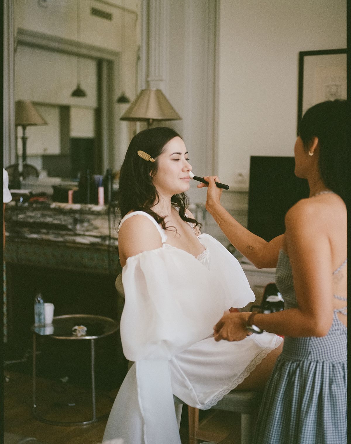 Carina in a white puff-sleeved robe having her makeup done at a dressing table