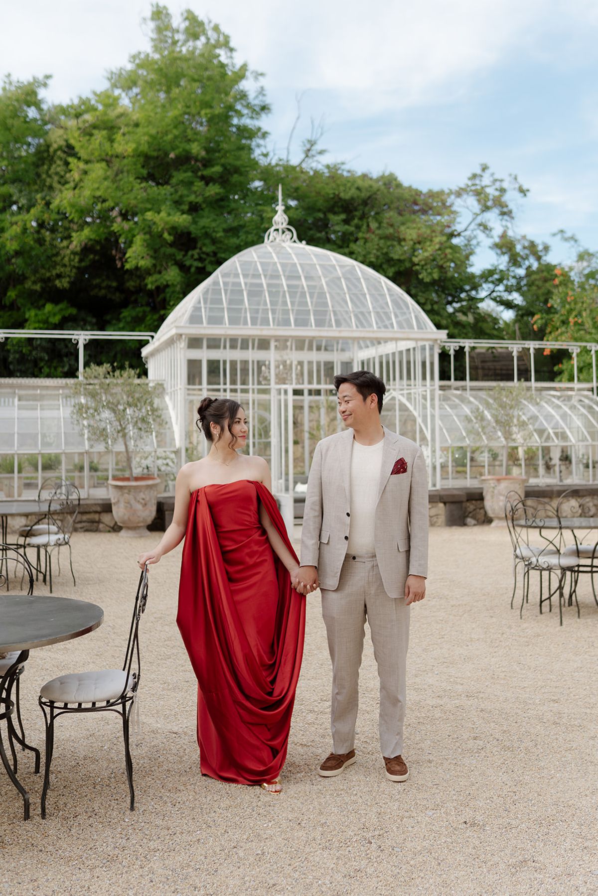 Carina in a draped red gown and Stefan in a linen suit holding hands by the greenhouse at the welcome party the day before
