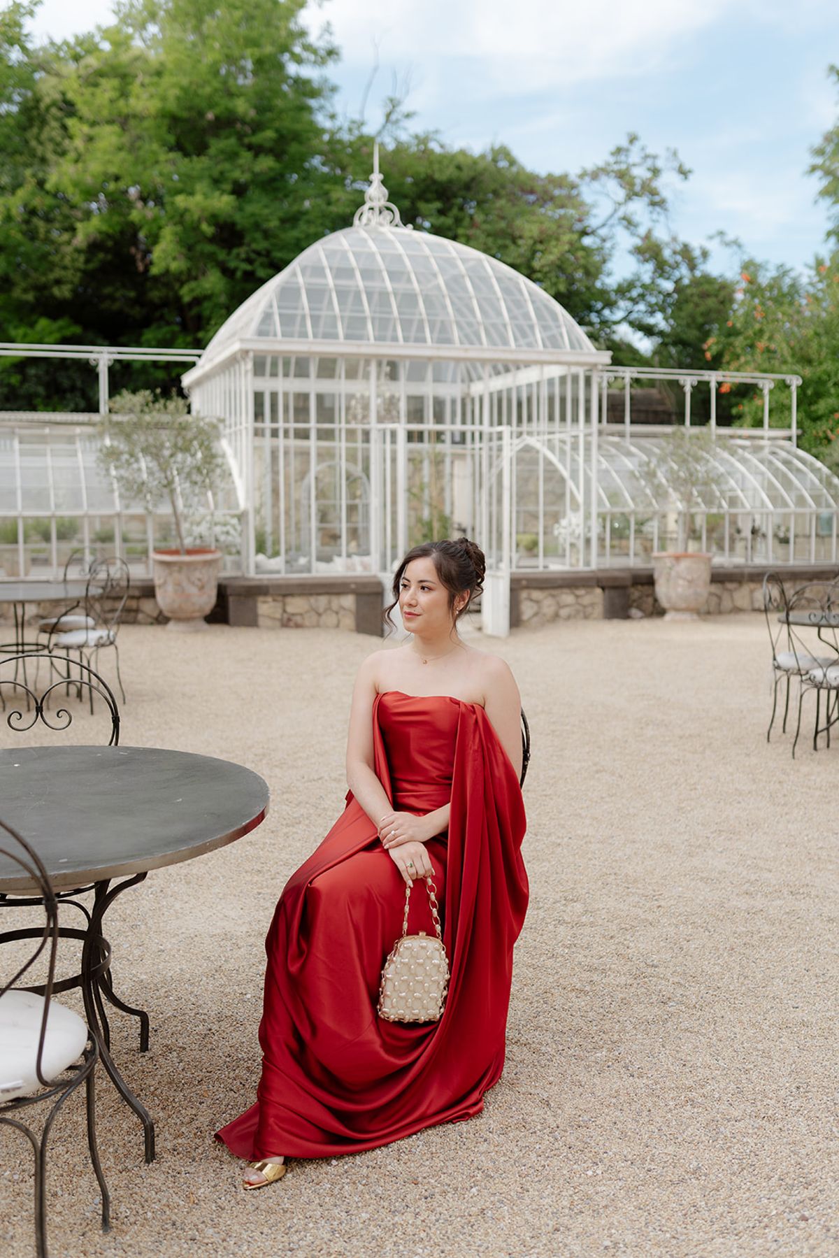 Carina in a draped red gown seated at a bistro table by the greenhouse at the welcome party the day before