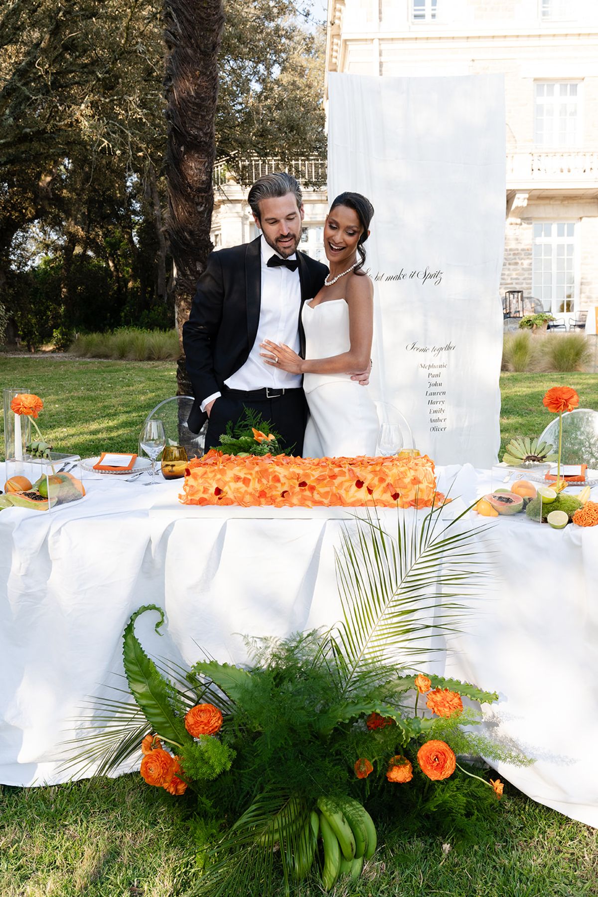 The bride and groom behind a table with a long cake covered in orange petals, the villa behind them