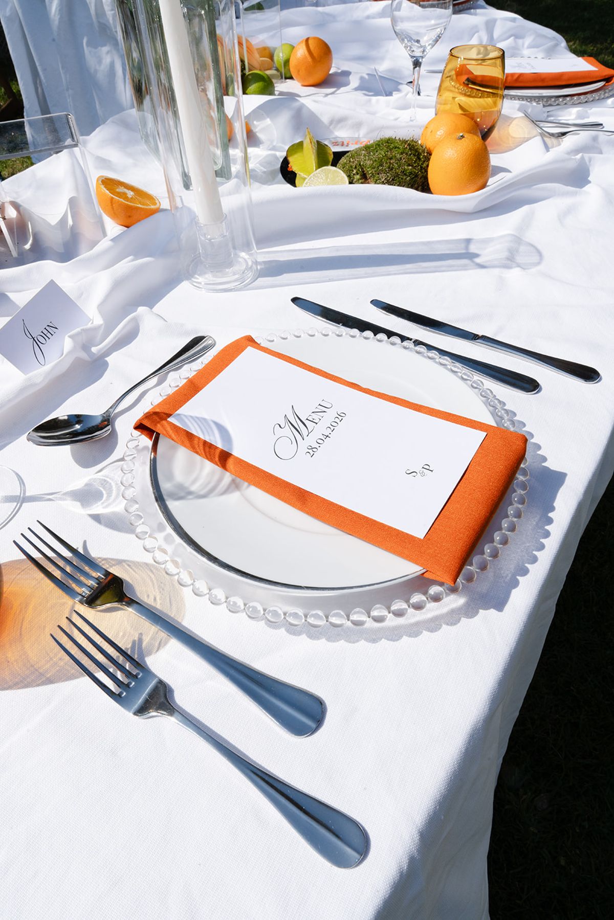 Place setting with an orange napkin, a printed menu and a name card on white linen, with citrus and candles