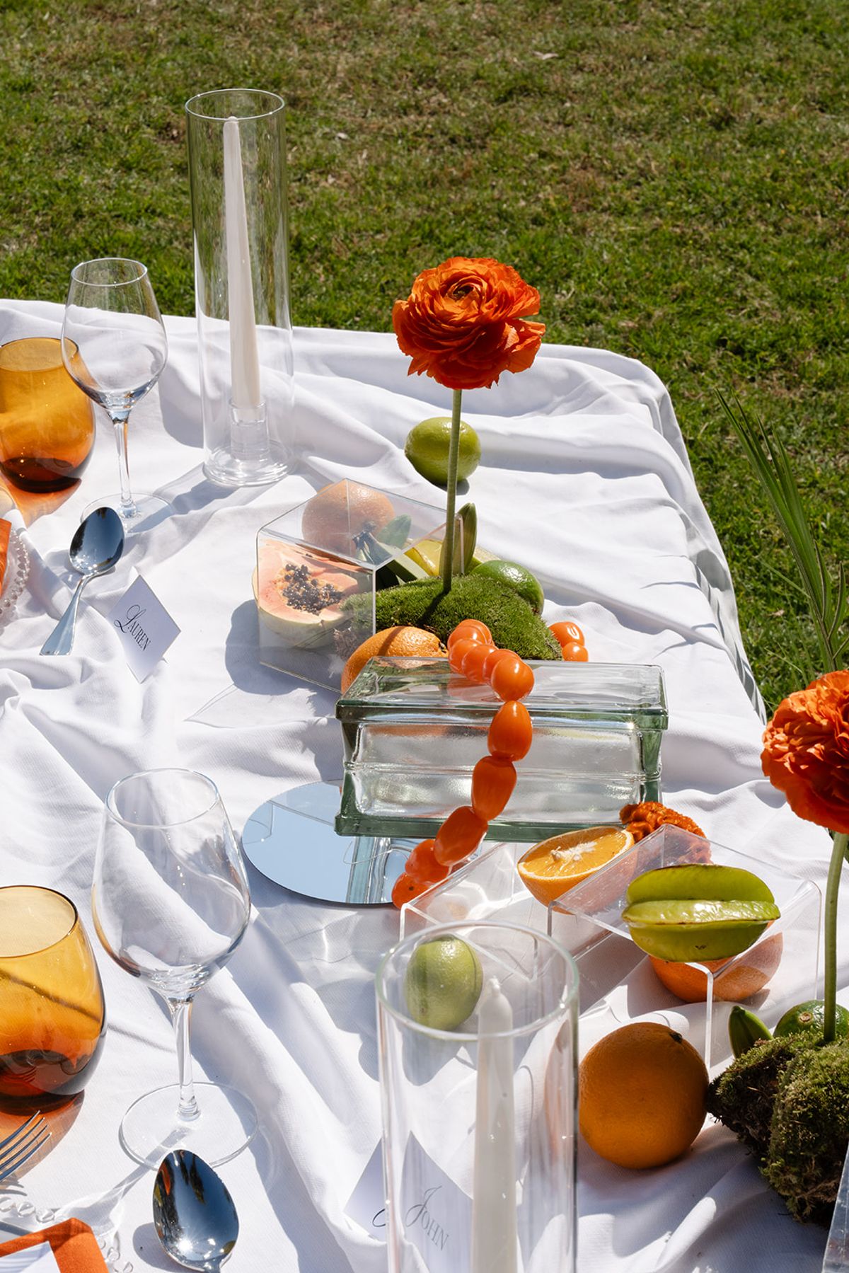 Centrepiece of an orange ranunculus in a glass cube with moss, citrus and clear glassware on white linen