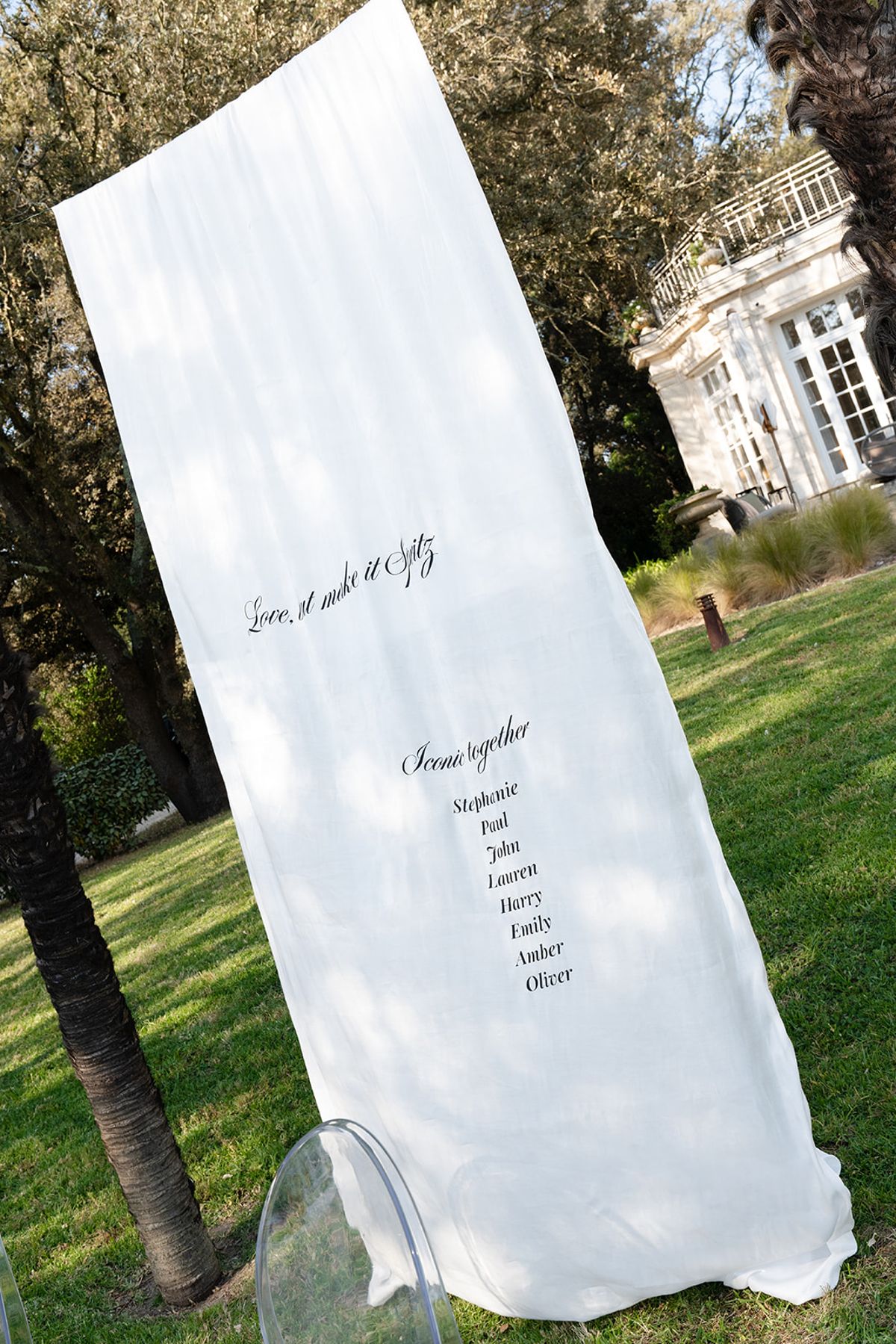 A white fabric seating banner lettered "Love, but make it Spritz" hanging from a tree on the lawn