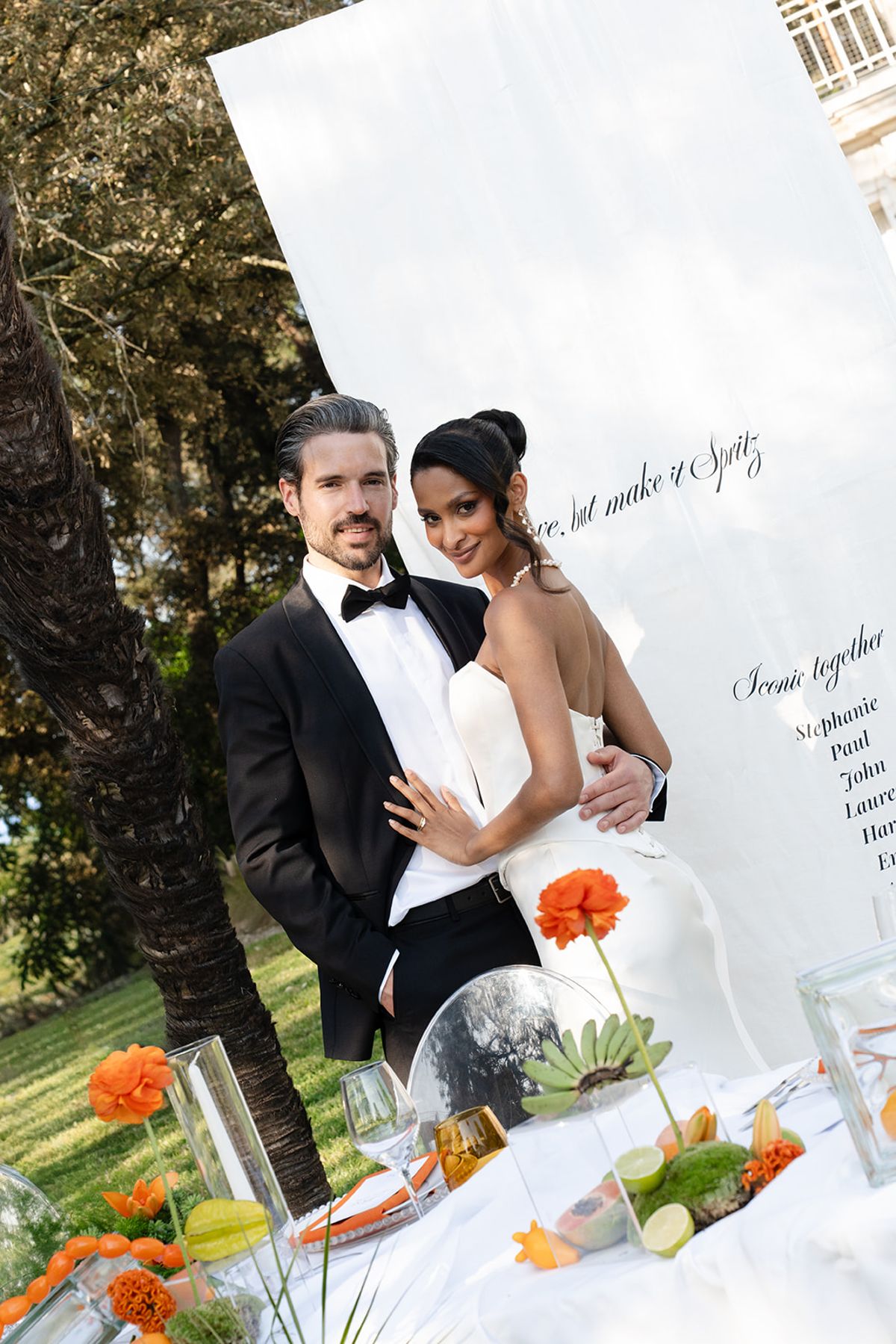 The groom with his arms around the bride beside a table of orange flowers and fruit, in front of a white fabric seating banner