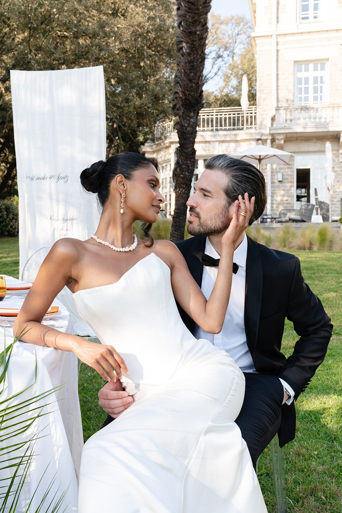 The bride seated on the groom's knee at the table on the lawn, her hand on his face