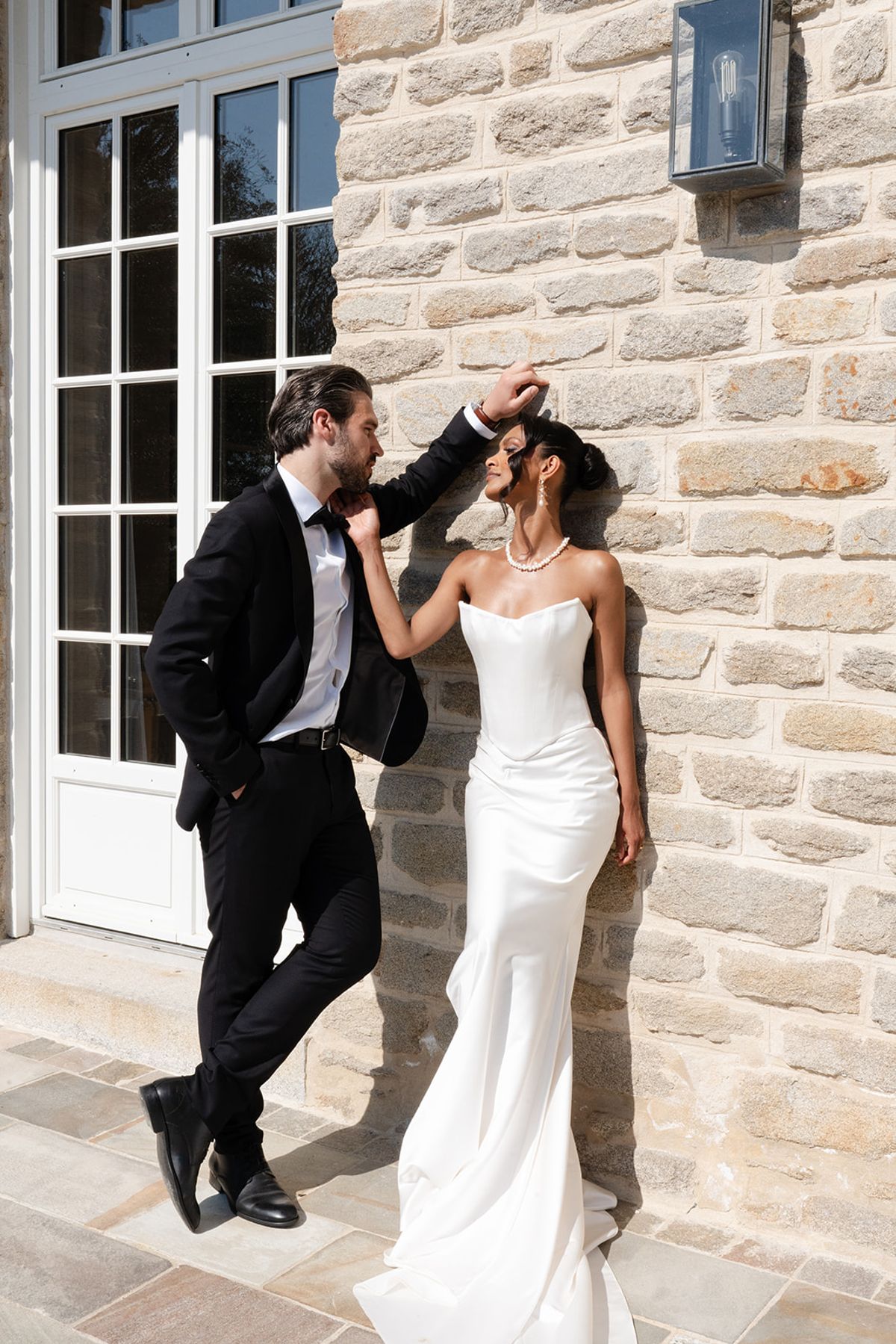 The groom leans over the bride against the villa's stone wall beside tall glazed doors