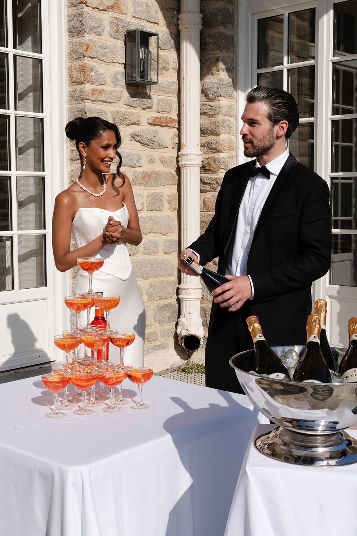 The groom pours champagne beside a tower of spritz coupes as the bride looks on, by the villa's stone wall