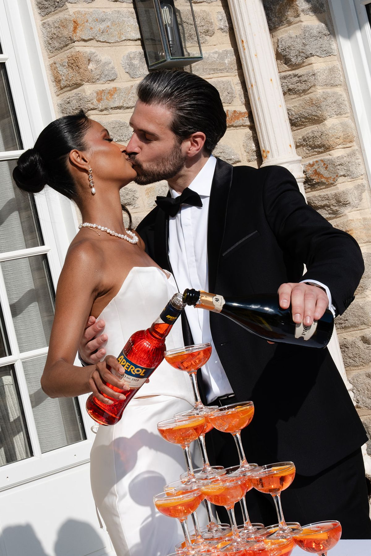 The bride and groom kiss as he pours champagne and she holds a bottle of Aperol over a tower of spritz coupes