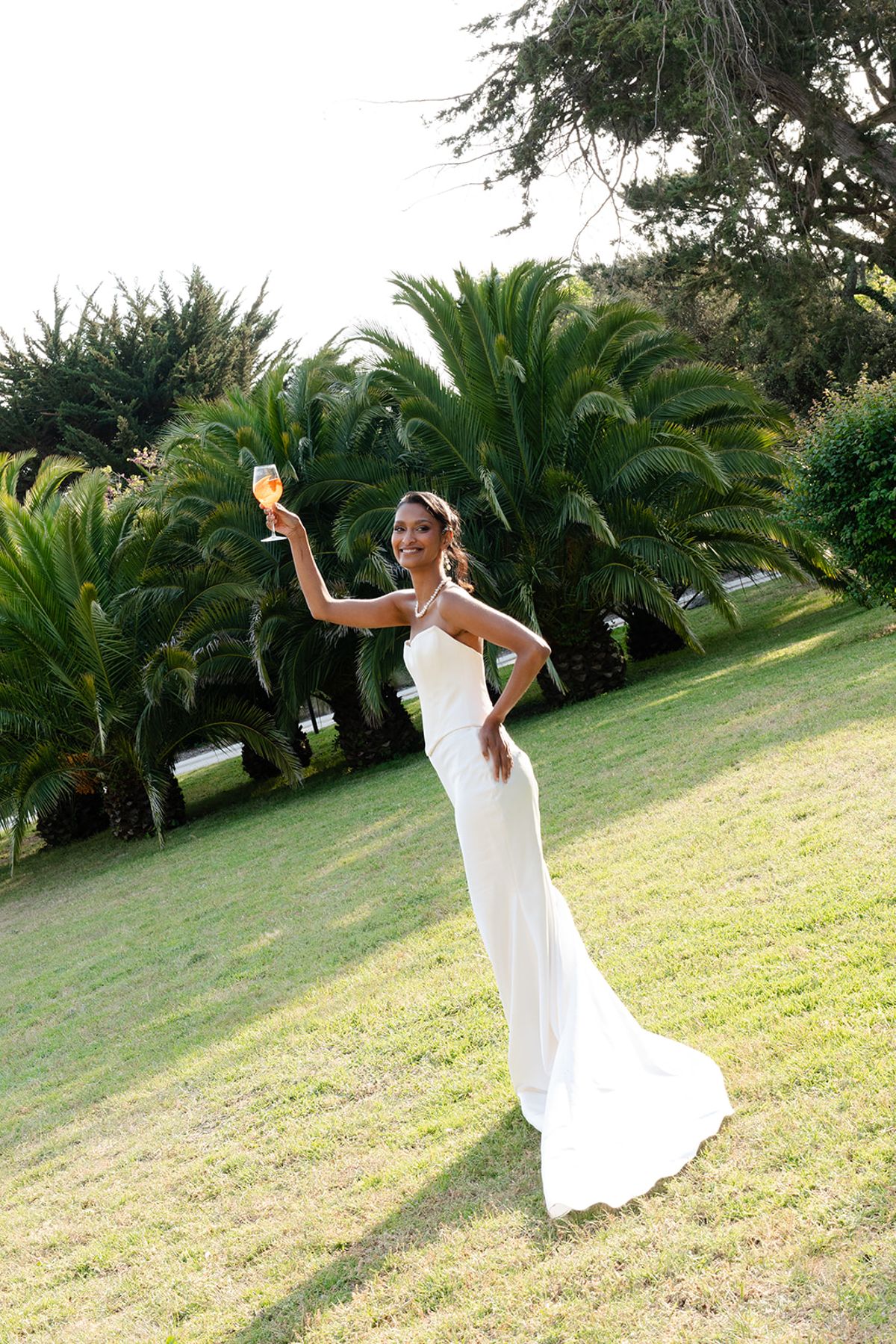 The bride raises a glass of spritz on the lawn, with palm trees behind her
