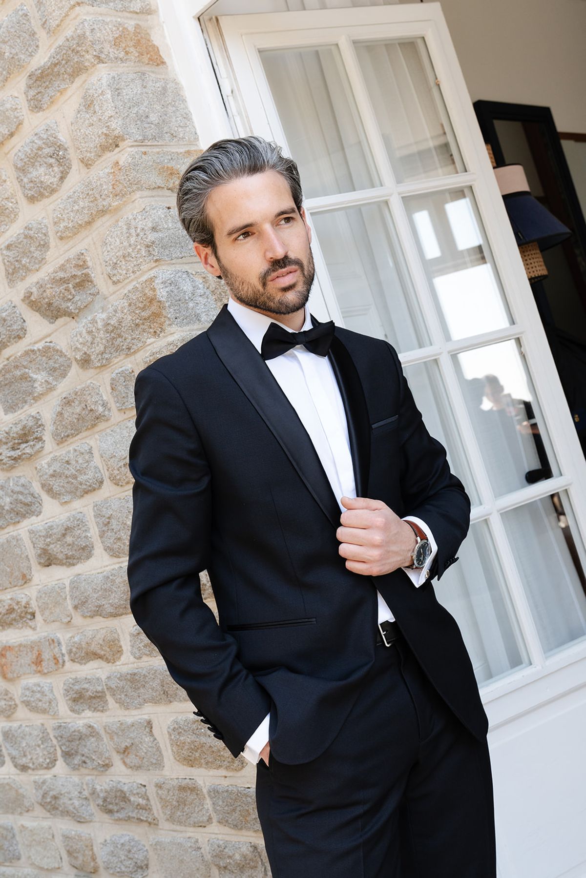 The groom in a tuxedo by tall glazed doors against the villa's stone wall