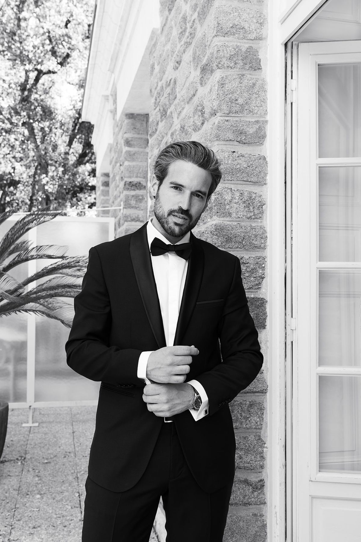 Black and white portrait of the groom in a tuxedo fastening his cuff against the villa's stone wall