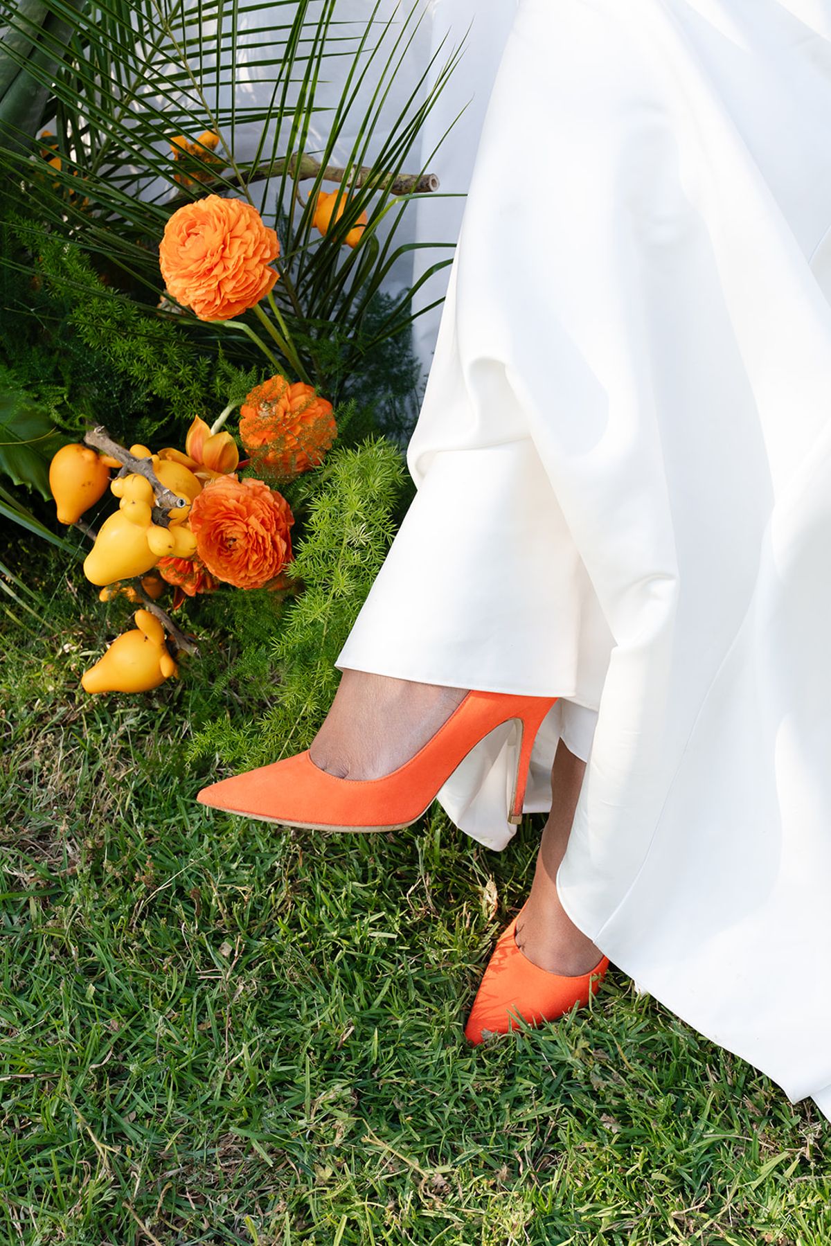 The bride's orange suede heel beneath her gown, beside orange ranunculus and small yellow fruit on the grass
