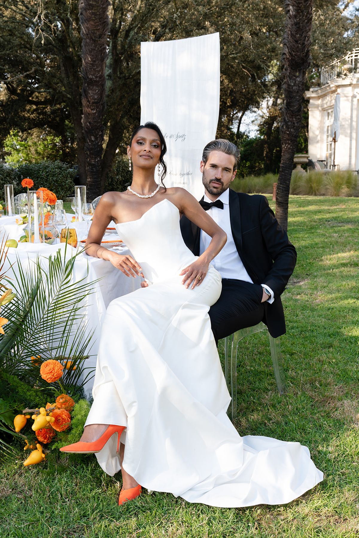 The bride, in orange heels, sits beside the groom at the table on the lawn