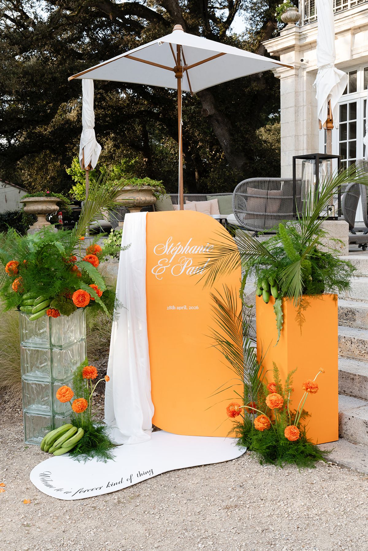 An orange welcome sign lettered for Stéphanie and Paul beneath a parasol, with orange flowers and palm leaves at the foot of the villa's steps
