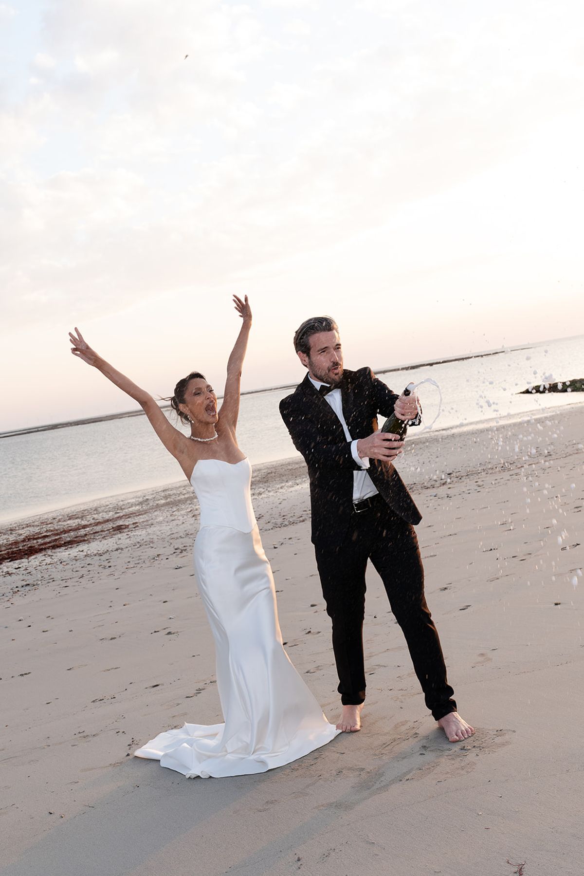 The bride throws her arms up as the barefoot groom sprays champagne on the beach at sunset
