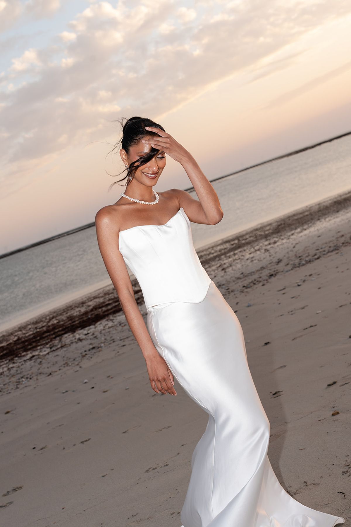 The bride in a strapless satin gown on the beach at sunset, one hand raised to her hair