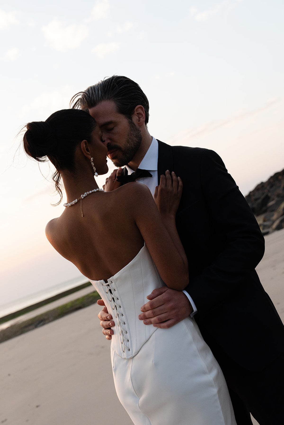 The bride and groom embrace on the beach at dusk, the lacing on the back of her gown visible