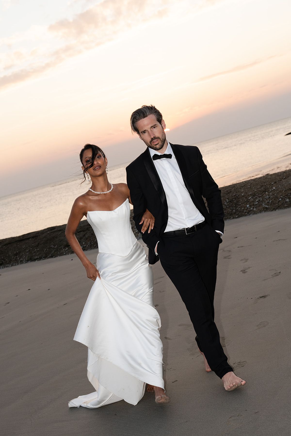 The bride and groom walk side by side along the beach at sunset