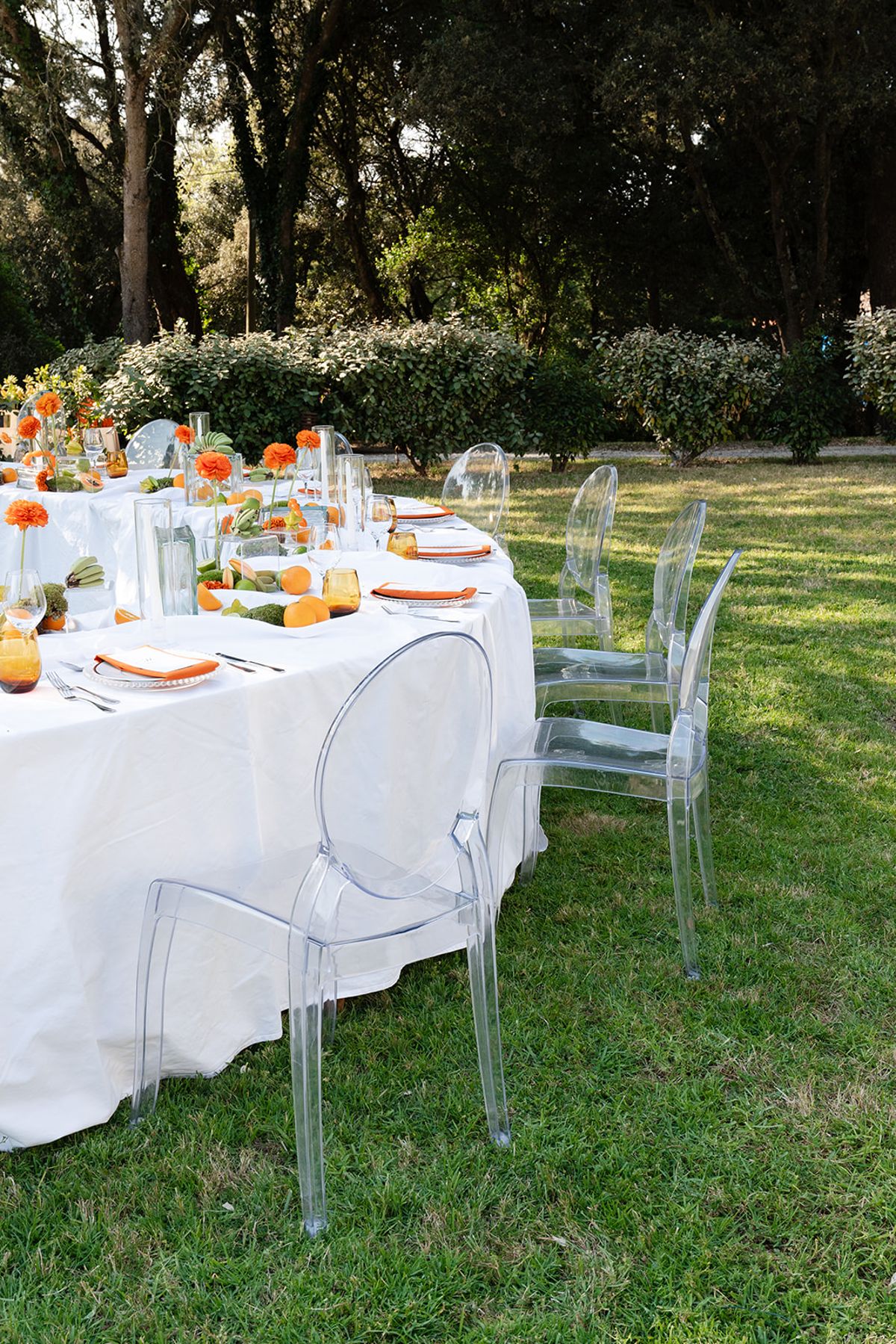 A round table dressed in white with clear chairs and orange flowers on the lawn beneath the trees
