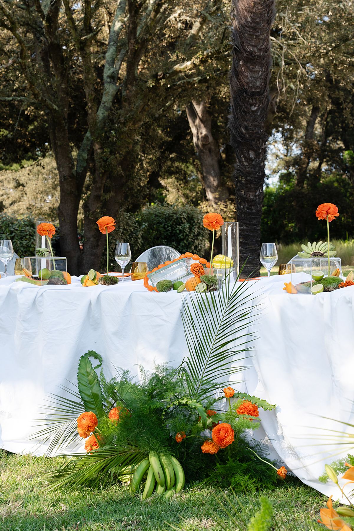 A long table dressed in white on the lawn, with palm leaves, orange flowers and bananas at its foot