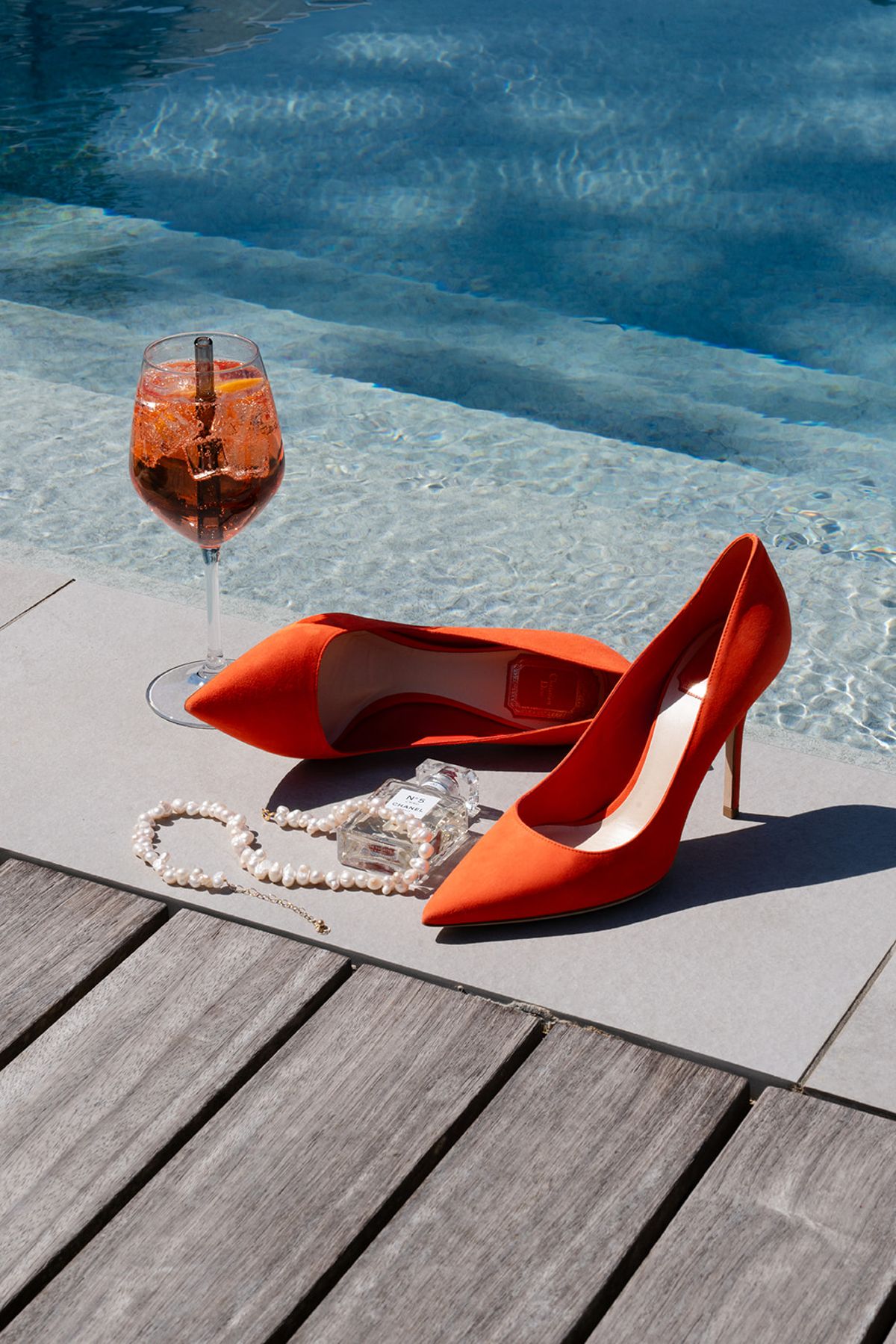 Orange suede heels, a pearl necklace and a glass of spritz at the edge of the swimming pool