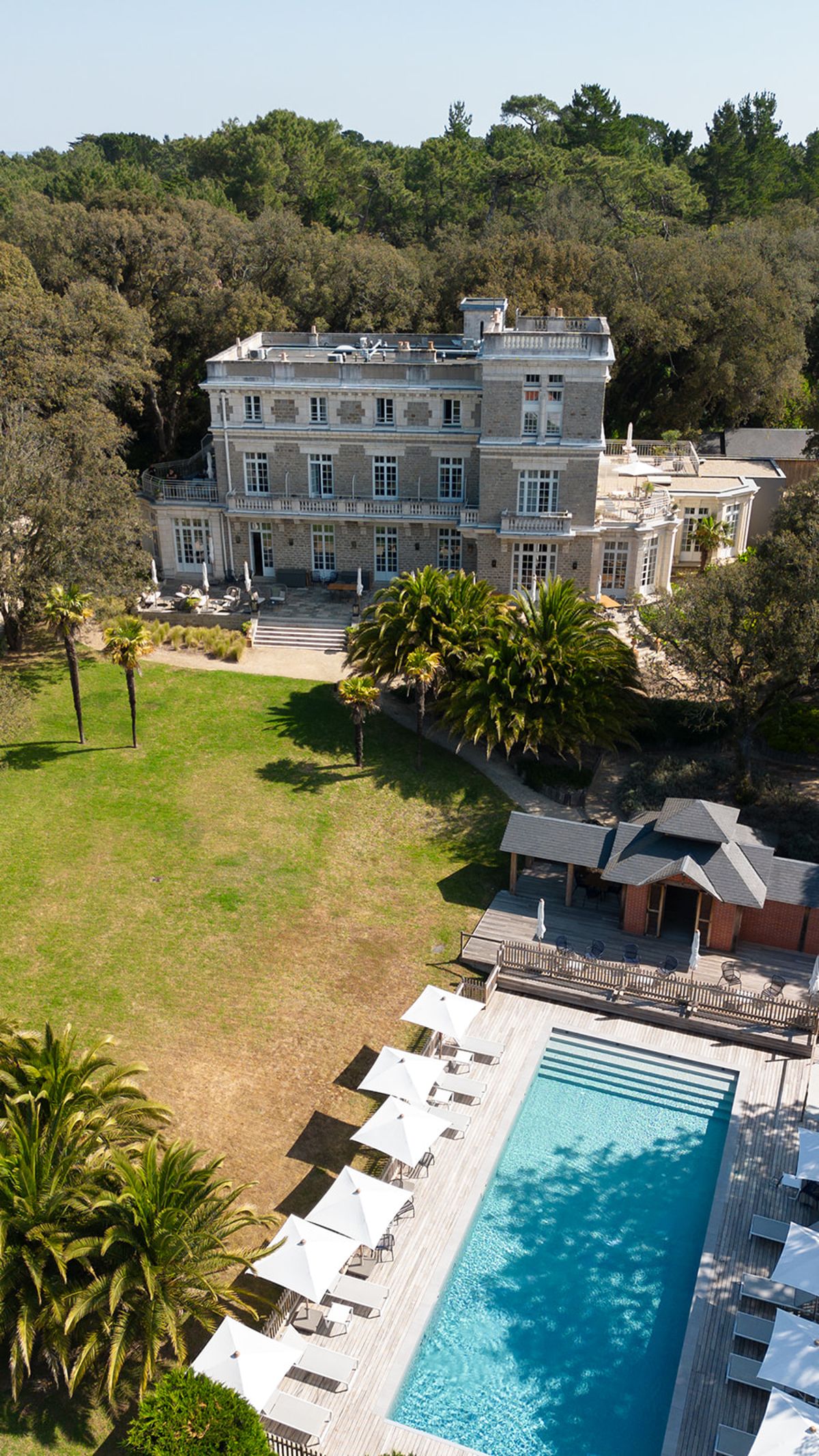Aerial view of the villa above its lawn, palm trees and swimming pool, surrounded by woodland