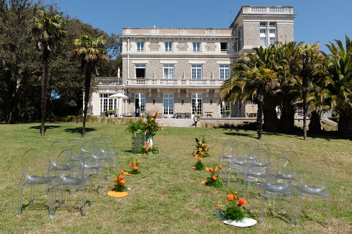 Ceremony set-up on the lawn, with rows of clear chairs and orange flowers along the aisle in front of the villa's stone façade and palm trees