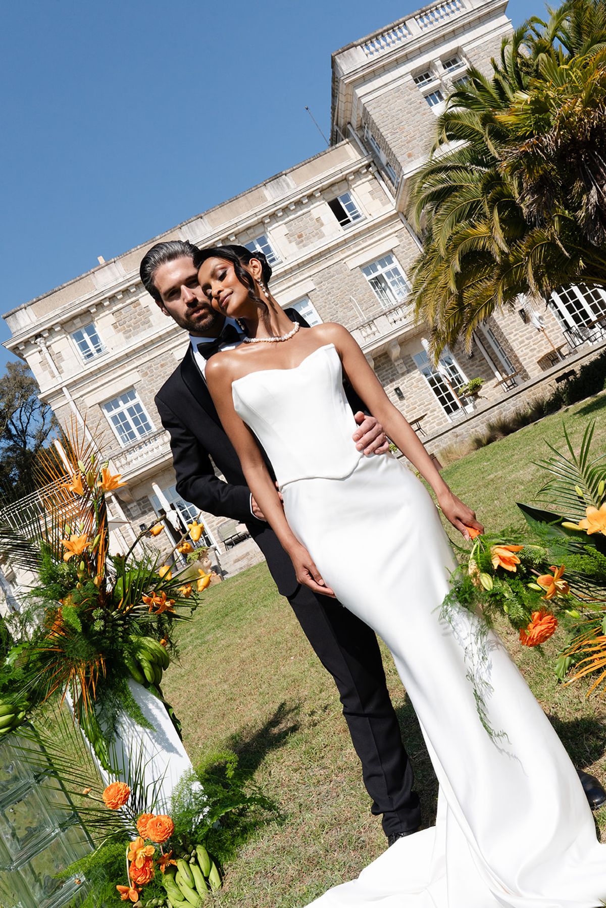 The groom holds the bride from behind between tropical floral arrangements in front of the villa's façade