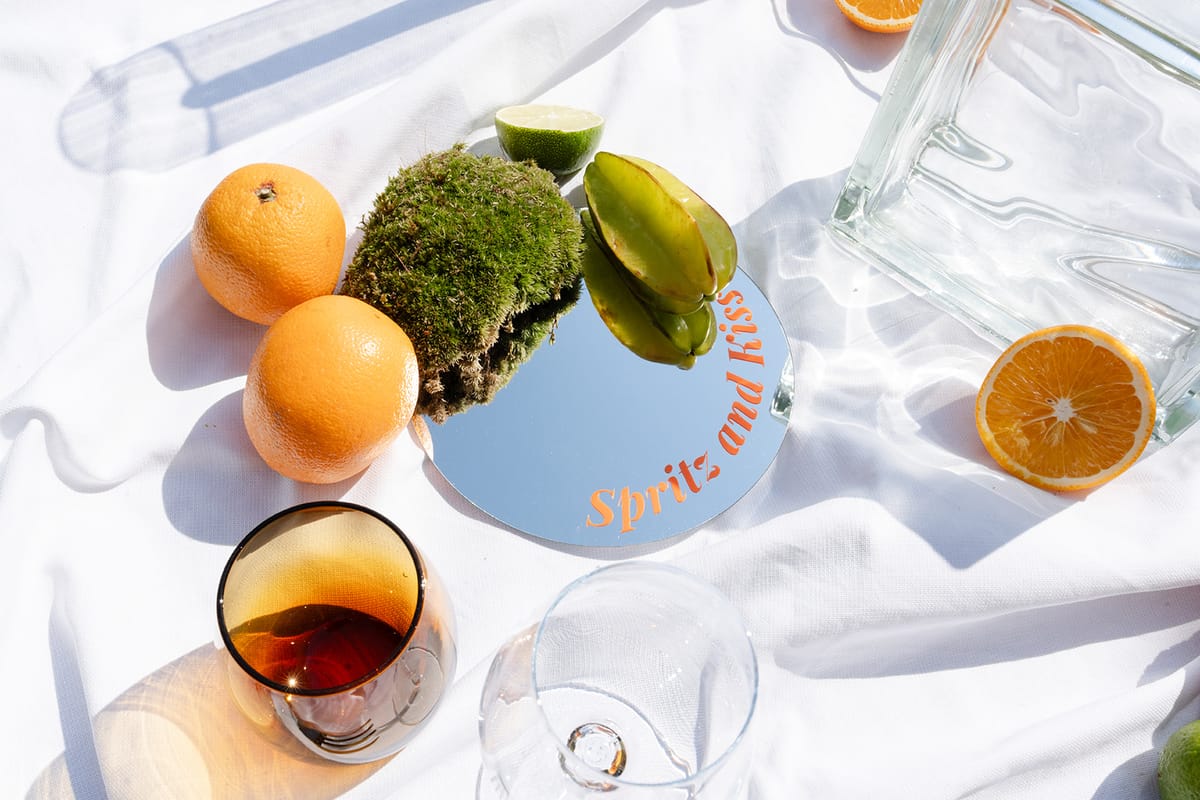 Oranges, a lime, moss and a mirrored coaster lettered in orange on white linen