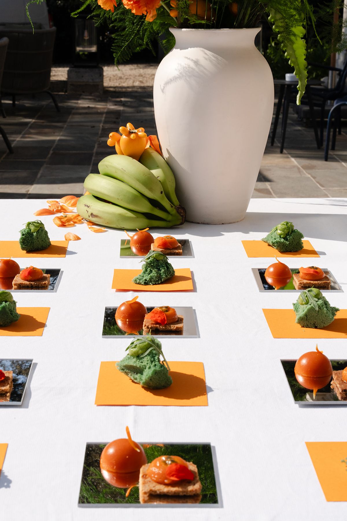 Rows of canapés on mirrored tiles and orange napkins, with a white vase and a bunch of green bananas