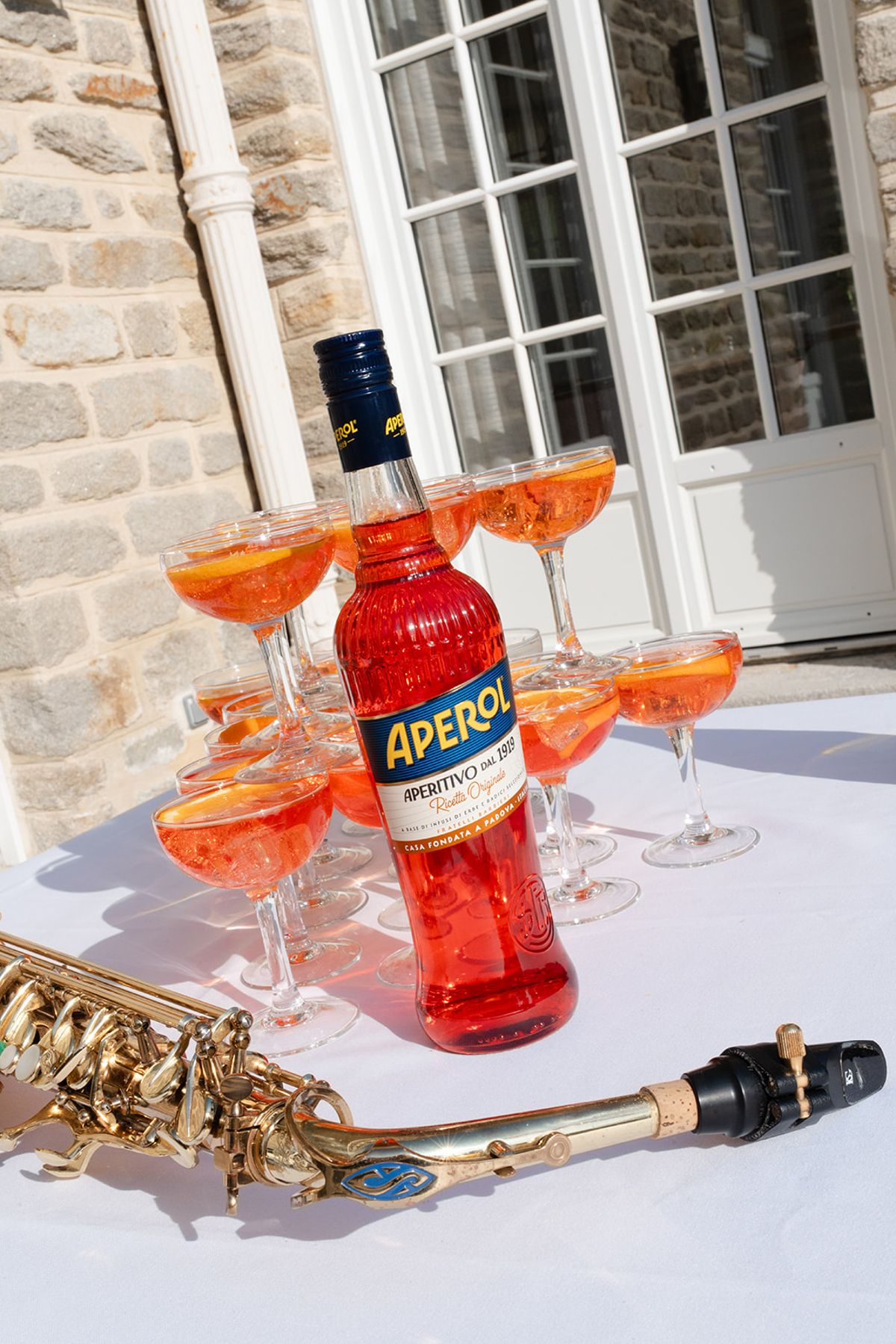 A bottle of Aperol, a tower of spritz coupes and a saxophone on a white table outside the villa