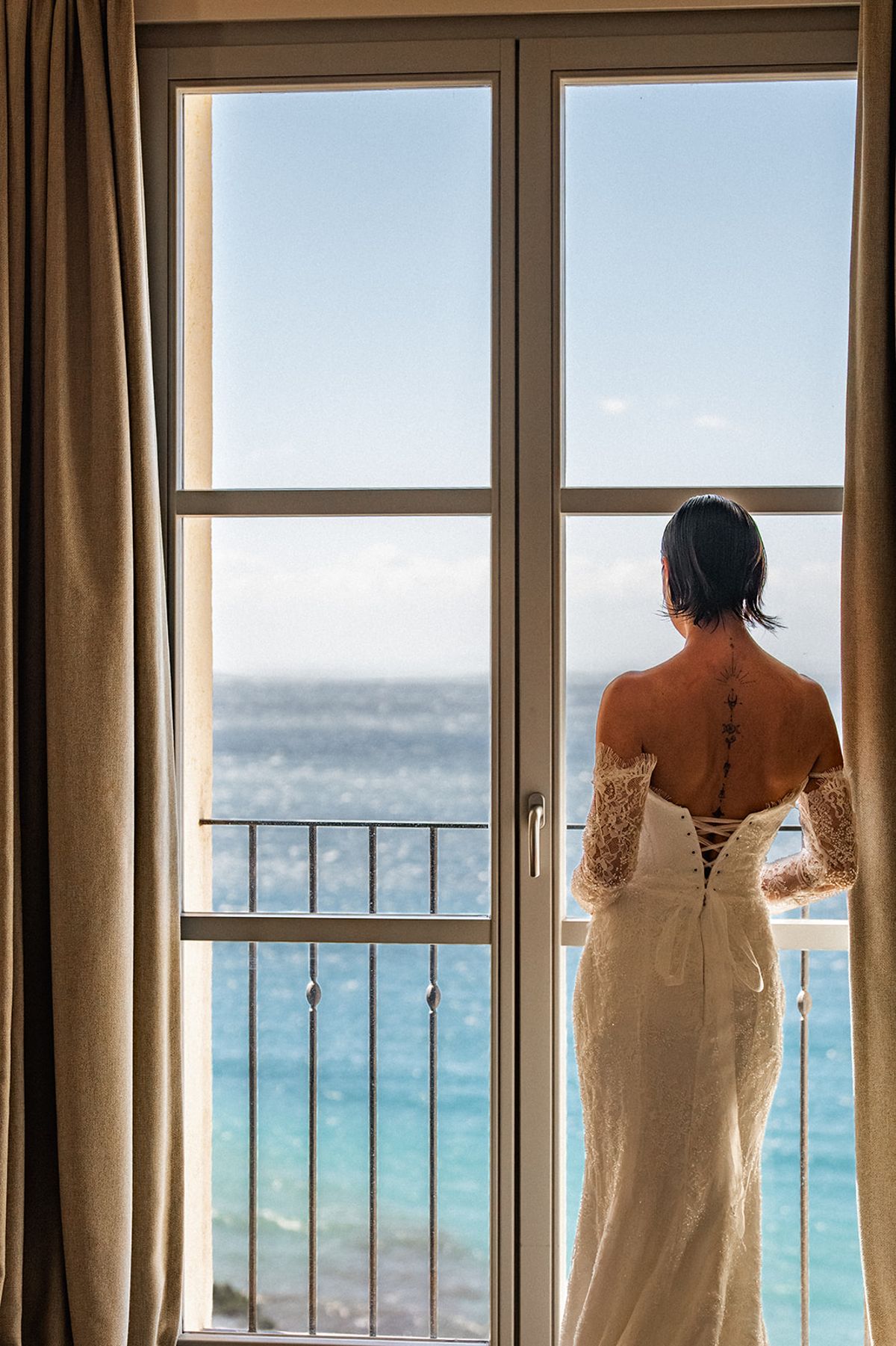 The bride stands at an open window in her gown, back to camera, looking out over the sea.