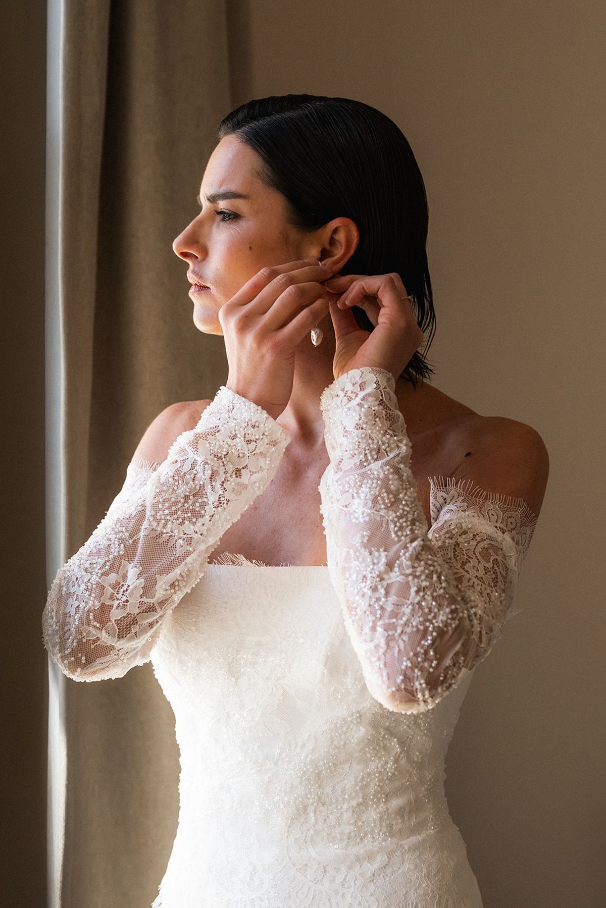 The bride fastens an earring, caught in profile in warm window light.