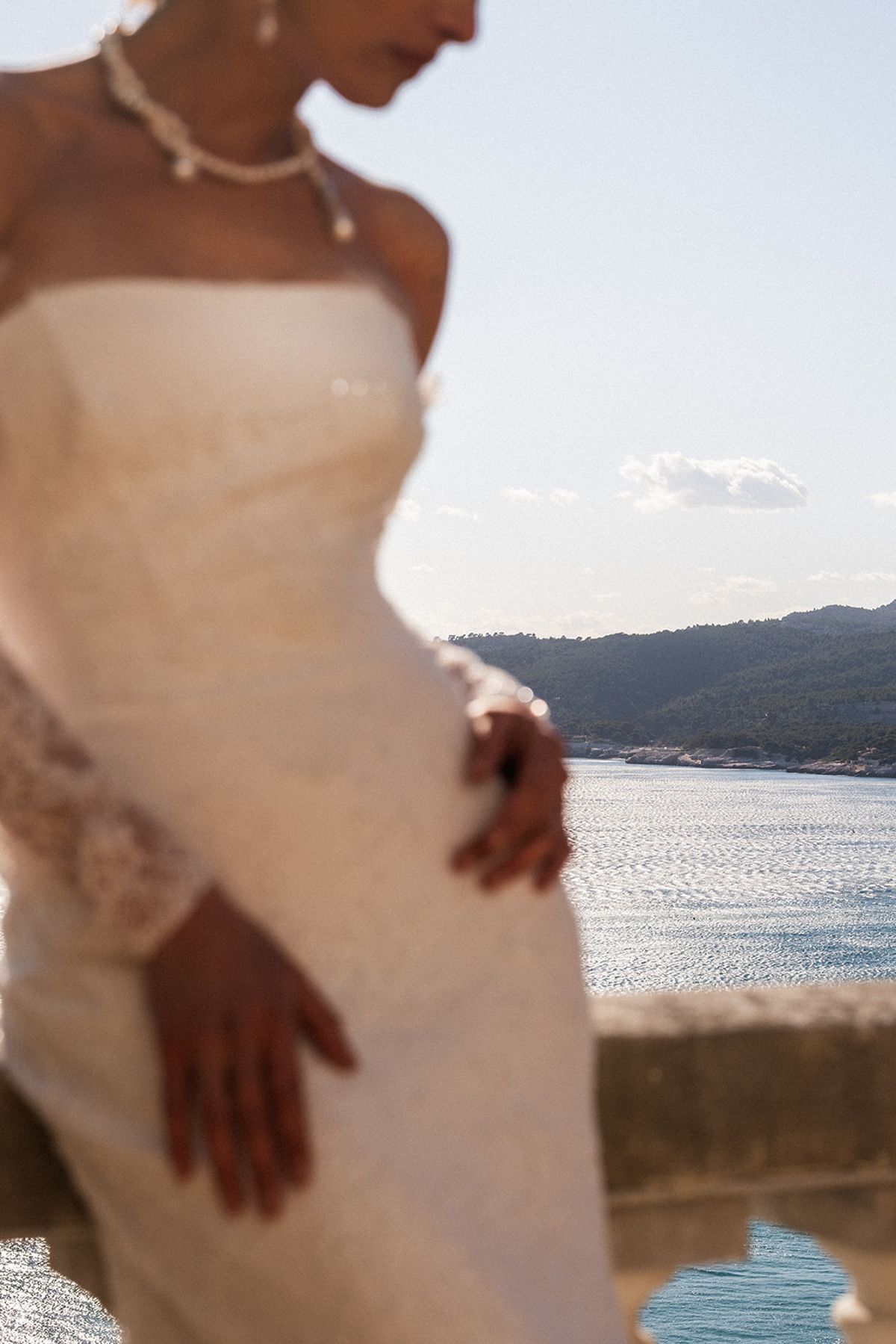 Close detail of the bride’s gown bodice and hand at her waist, the Mediterranean coastline soft-focused behind her.