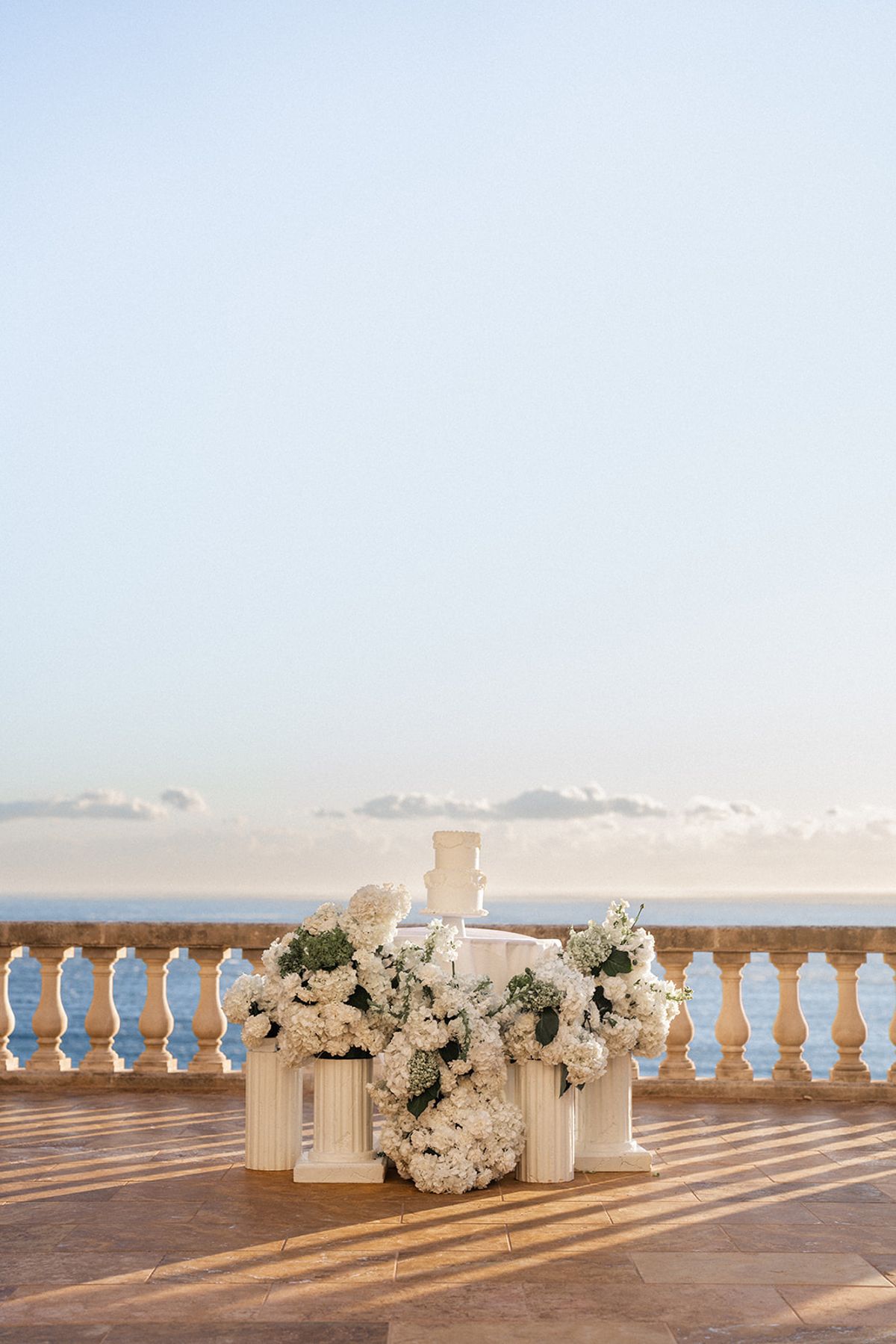 A two-tier white wedding cake sits on a pedestal table banked with white hydrangea, the sea visible beyond the terrace railing.